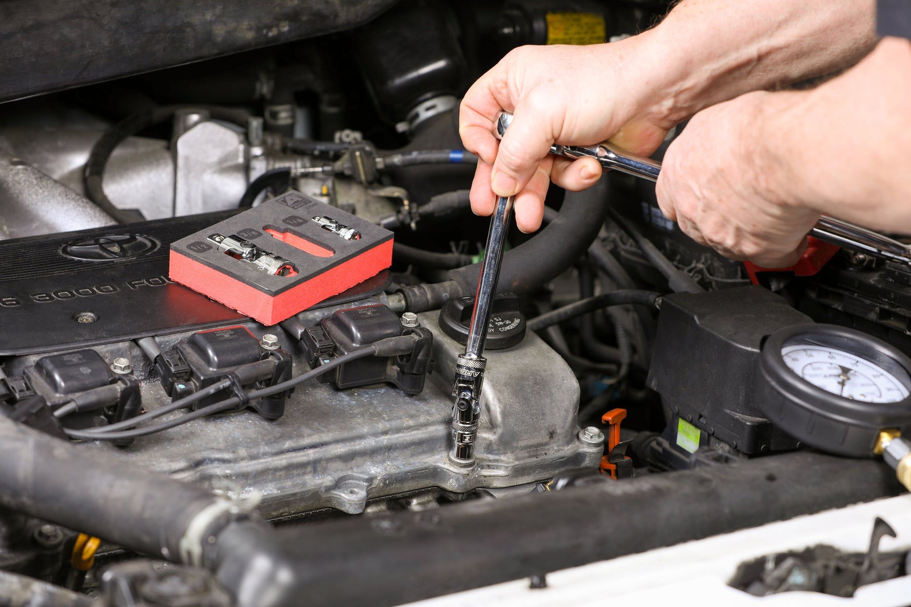 A set of universal joint sockets sits on an open engine bay, showcasing two universal joint sockets. A man out of frame uses a universal joint socket and ratchet to unscrew a fastener inside of the engine bay.