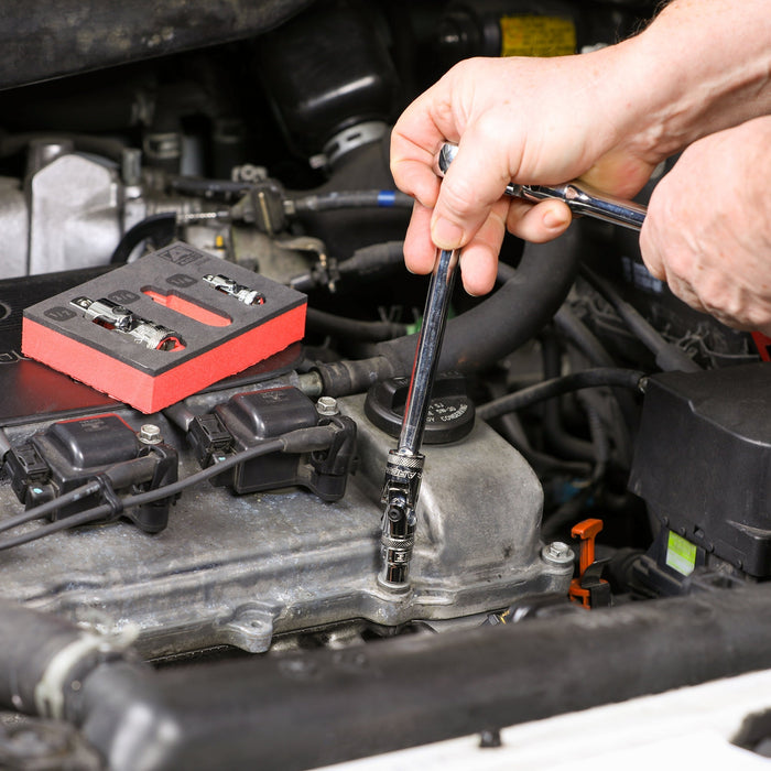 A set of universal joint sockets sits on an open engine bay, showcasing two universal joint sockets. A man out of frame uses a universal joint socket and ratchet to unscrew a fastener inside of the engine bay.